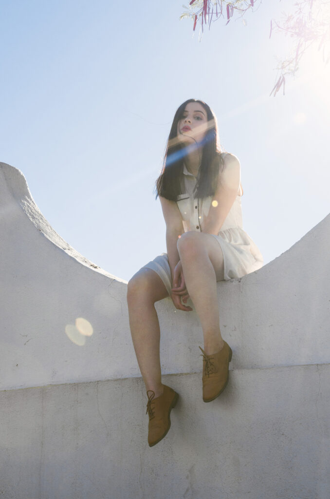 Gabriela Inzunza sits atop a white curved structure bathed in sunlight with a tree branch bearing red leaves overhead.